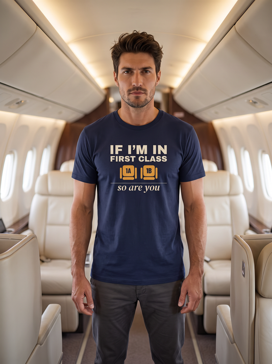 Liora standing in the aisle of a first-class aircraft cabin, wearing the unisex t-shirt in navy with two wide seats marked 1A and 1B in amber and cream type reading if I'm in first class, so are you. Model: Liora (AI brand character). AI-generated model image; the printed design is the actual product artwork.