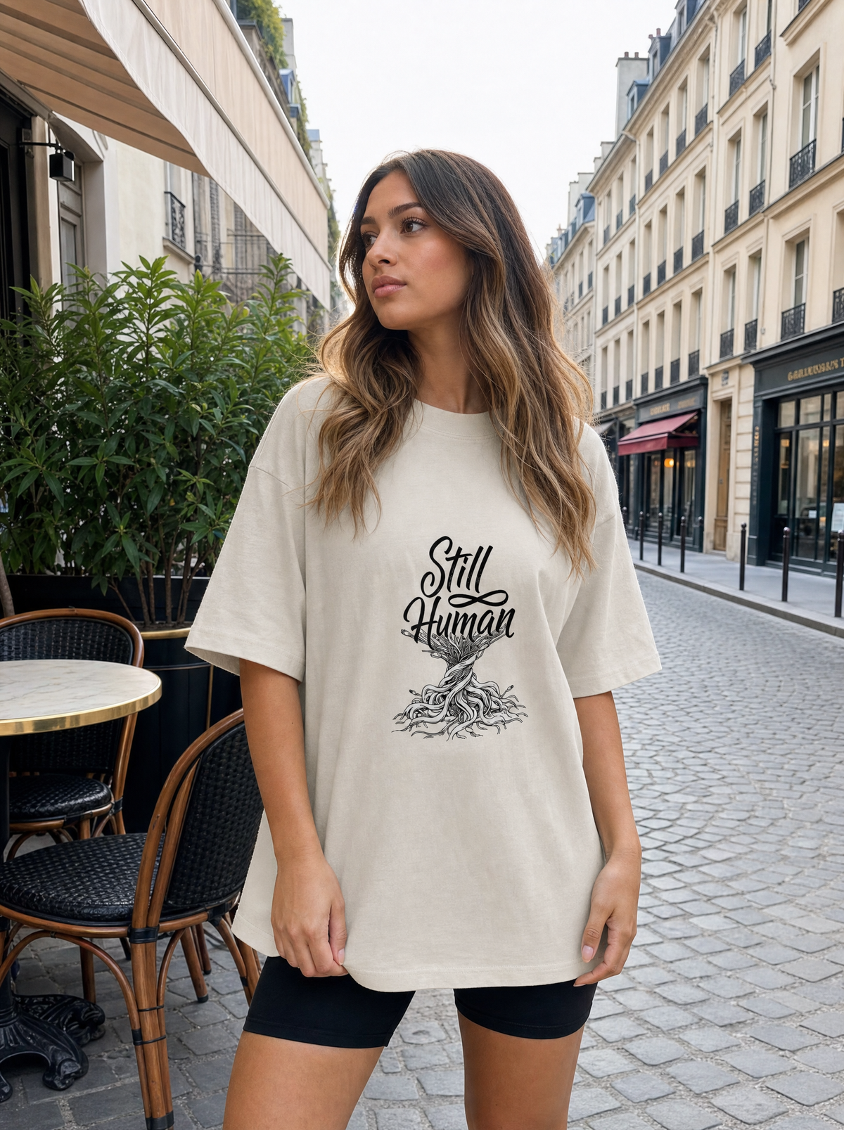 Still Human oversized faded tee in bone, worn on a city street by Seloura, Printelier's female brand character. AI-generated model image; the printed design is the actual product artwork.