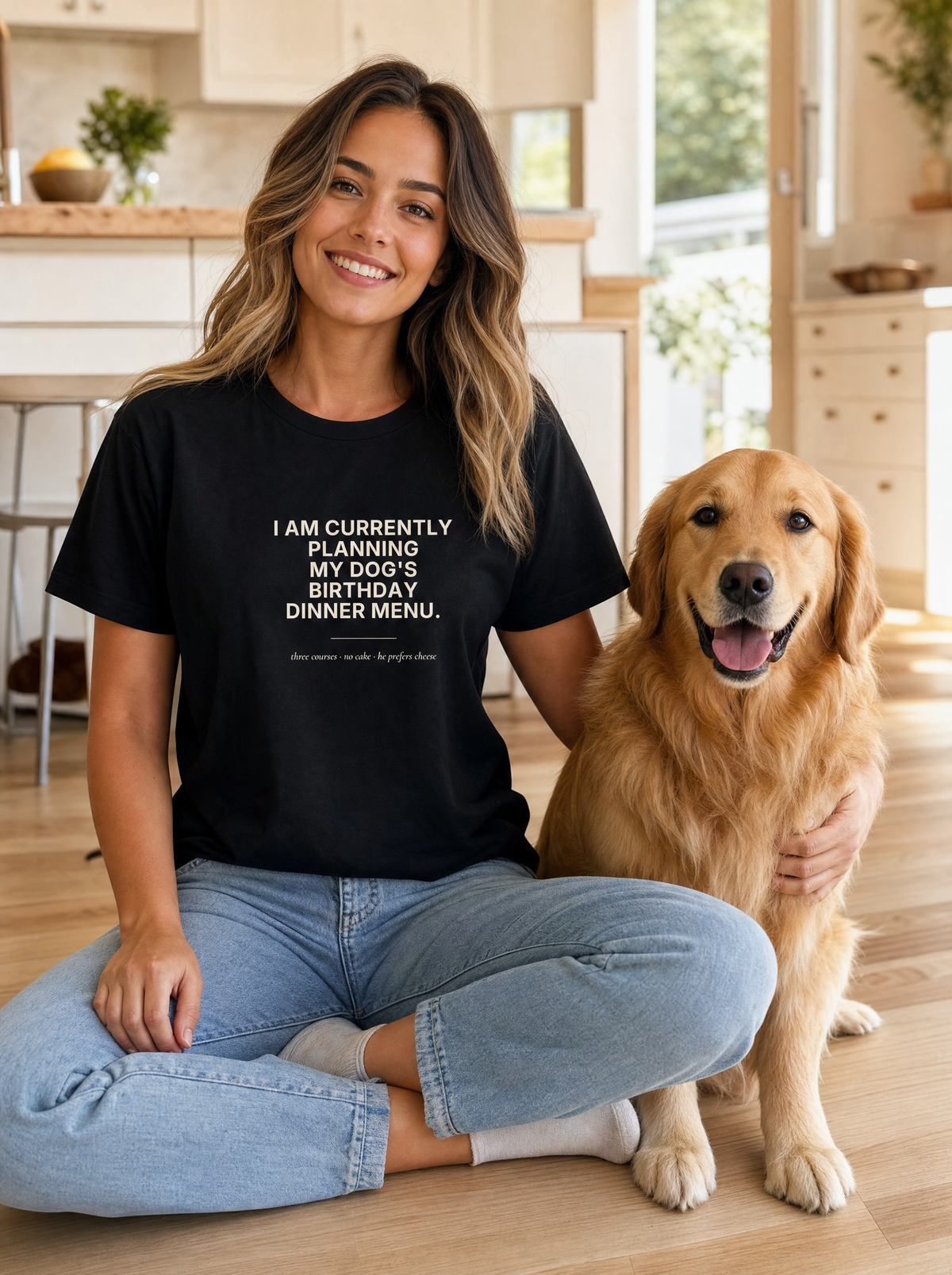 Seloura sitting on a kitchen floor next to a golden retriever, wearing the black My Dog's Birthday Dinner Menu tee with cream text. Model: Seloura (AI brand character). AI-generated model image; the printed design is the actual product artwork.