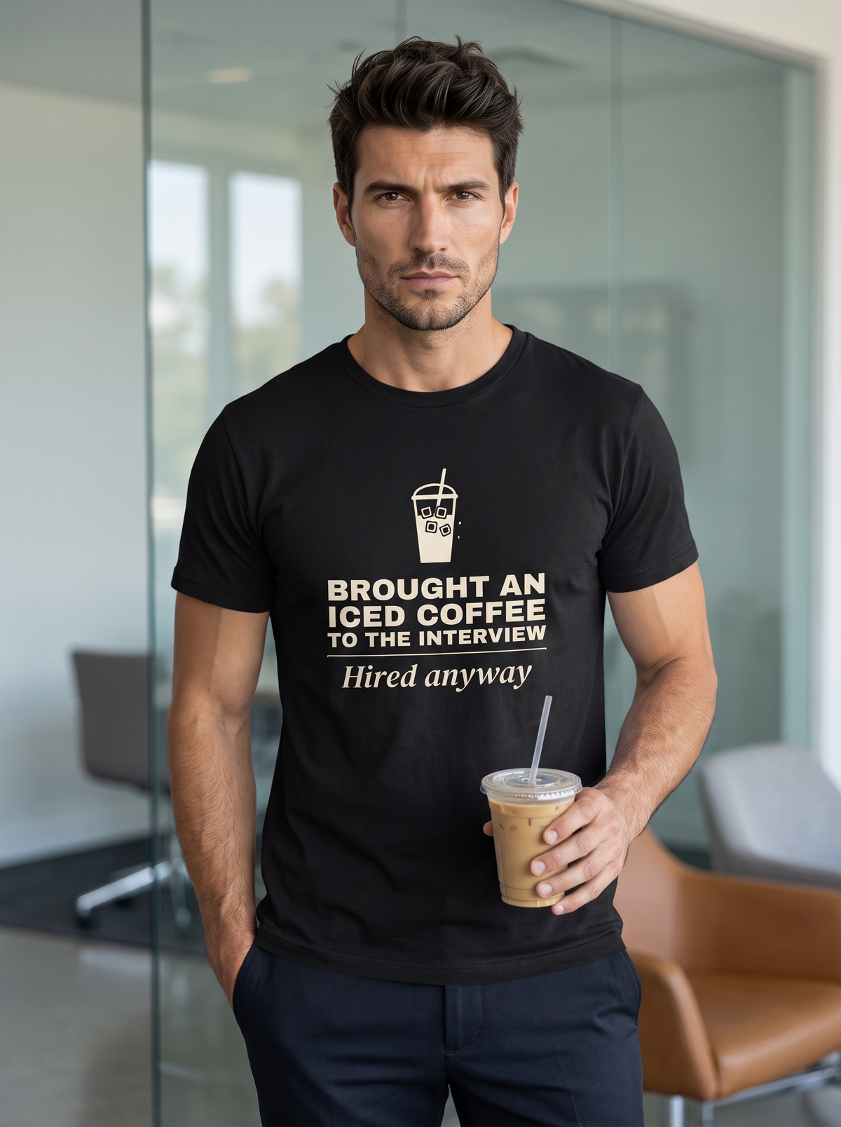 Liora standing in an office reception holding an iced coffee, wearing the unisex t-shirt in black with a drawing of an iced coffee cup and cream bold type reading brought an iced coffee to the interview, hired anyway. Model: Liora (AI brand character). AI-generated model image; the printed design is the actual product artwork.