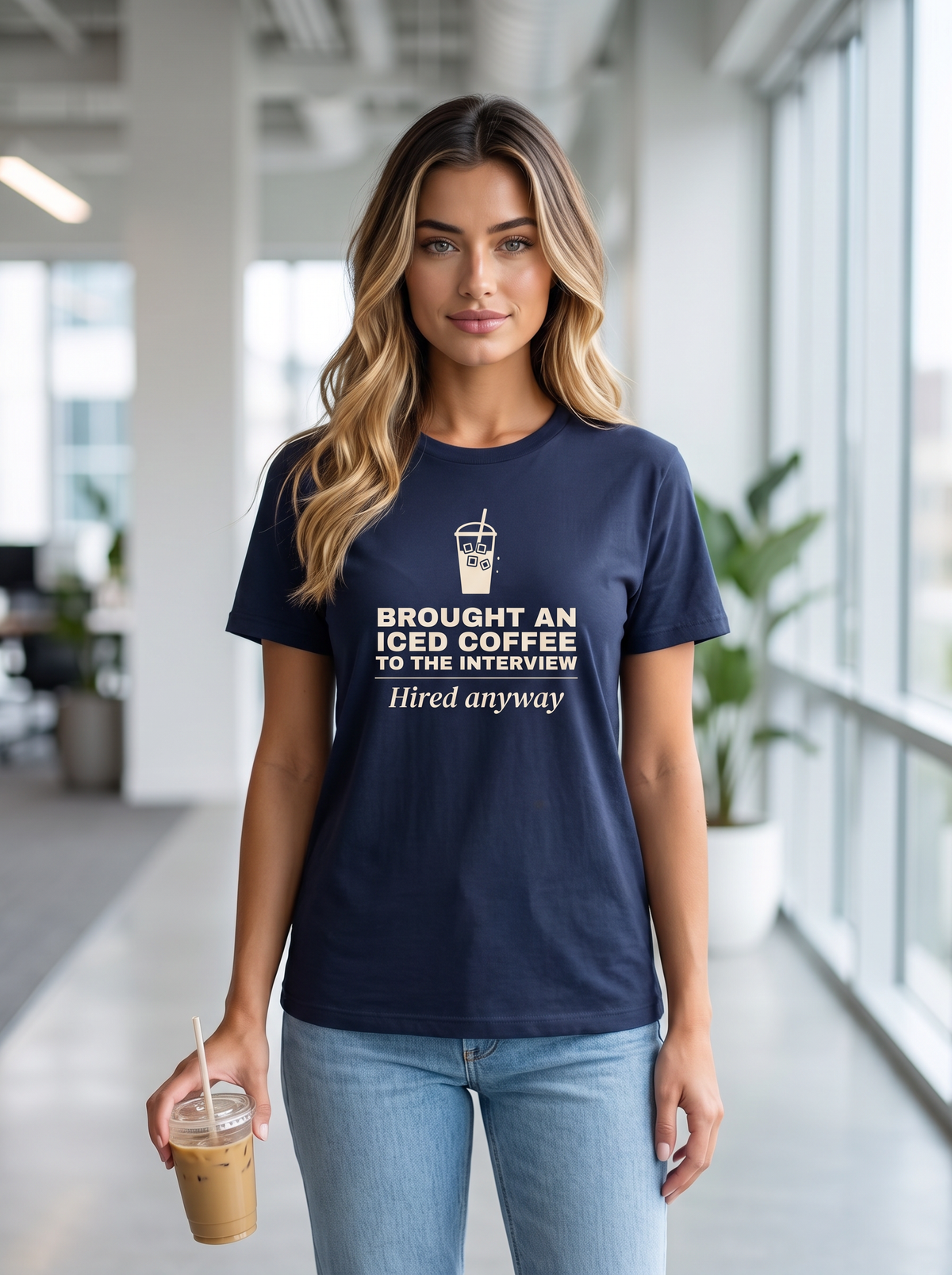 Seloura standing in an office corridor holding an iced coffee, wearing the unisex t-shirt in navy with a drawing of an iced coffee cup and cream bold type reading brought an iced coffee to the interview, hired anyway. Model: Seloura (AI brand character). AI-generated model image; the printed design is the actual product artwork.