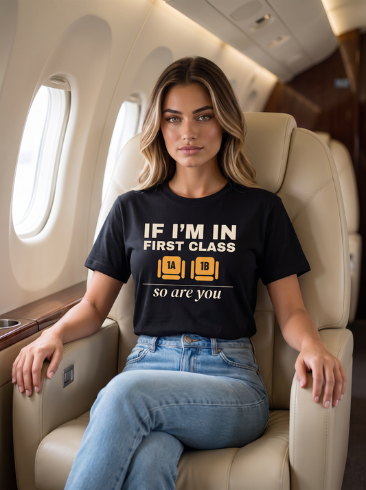 Seloura sitting in a wide first-class aircraft seat, wearing the unisex t-shirt in black with two wide seats marked 1A and 1B in amber and cream type reading if I'm in first class, so are you. Model: Seloura (AI brand character). AI-generated model image; the printed design is the actual product artwork.