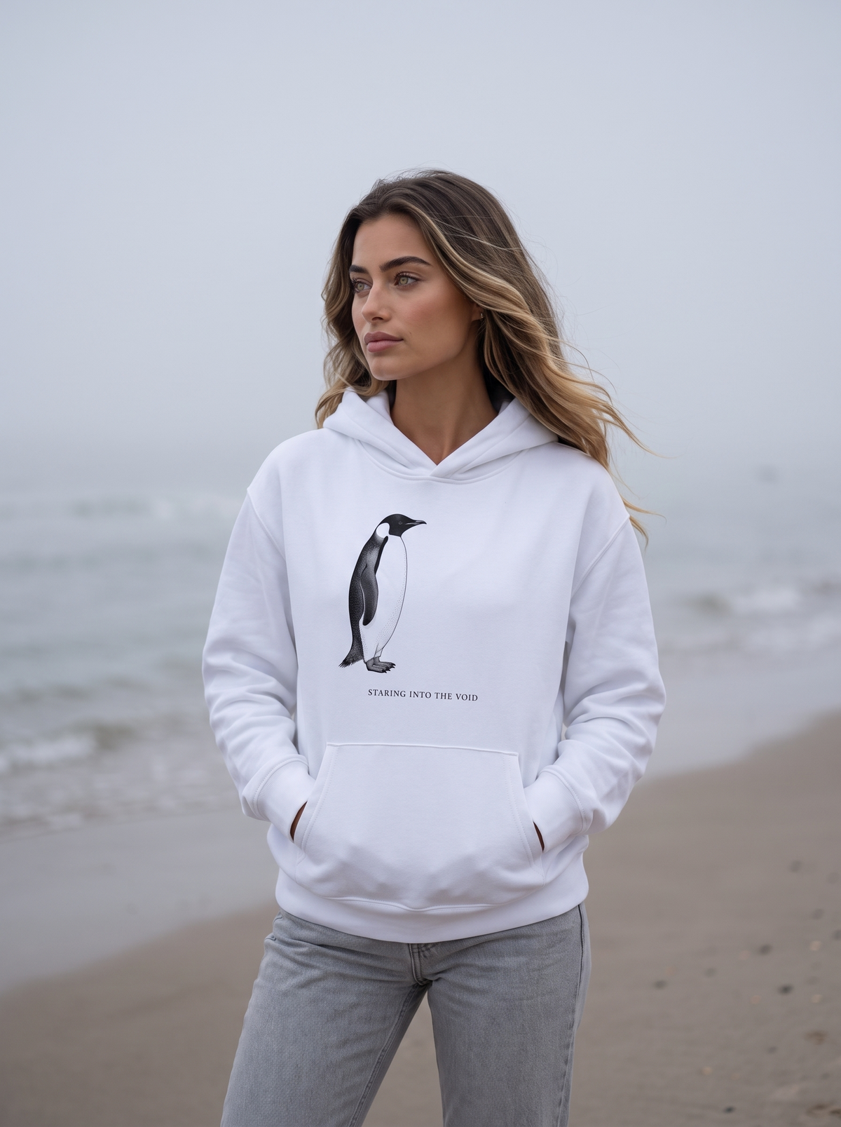 Seloura standing on an empty misty beach with her hands in the pocket, looking out to sea, wearing the oversized heavyweight hoodie in white with a vintage engraving of a penguin and the line staring into the void. Model: Seloura (AI brand character). AI-generated model image; the printed design is the actual product artwork.